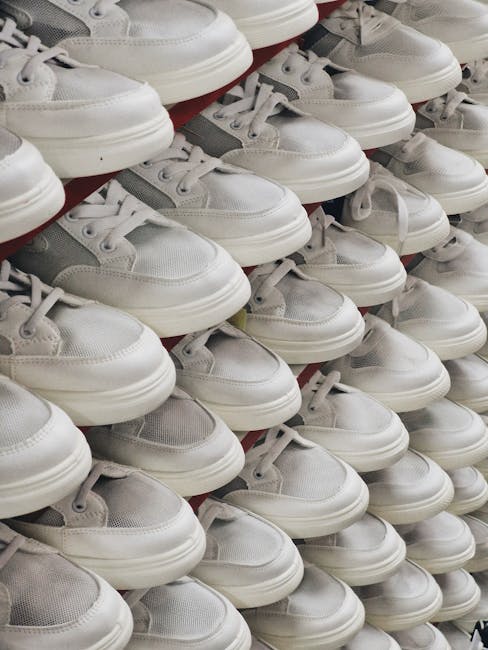 A large collection of white and grey sneakers arranged in neat rows on a display shelf, with their toes pointing forward and laces tied. The shoes appear to be made of leather and mesh materials, featuring a smooth, shiny finish on the white sections and matte grey accents. The sneakers are stacked closely together, occupying the entire visible surface of the shelf, which is situated in a retail environment with bright, even lighting. The background shows a blurred view of other shoes and store fixtures, indicating an indoor setting dedicated to footwear display. This image relates to private or alternative disposal options, such as donations or resale, which can be part of rubbish removal or waste management services provided by companies like Waste Disposal Hackney, especially when managing large quantities of footwear destined for bulk collection or recycling.