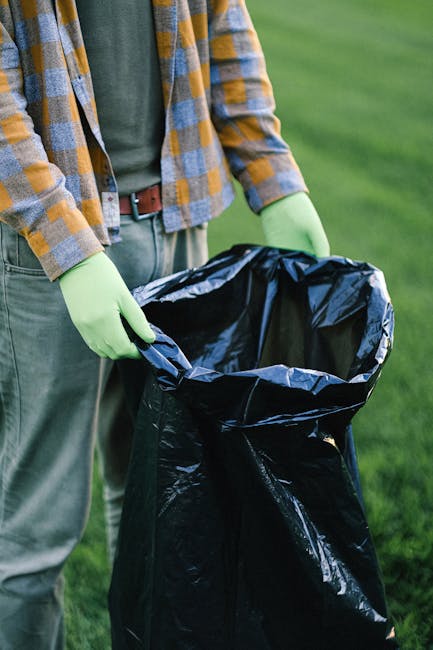 Three large black plastic rubbish sacks are positioned along the curbside on a paved street, with some bags appearing to be partially torn or crumpled, revealing mixed waste inside. The sacks are situated close to a black metal fence with vertical bars, which runs parallel to the pavement and encloses a garden area with dense foliage and trees in the background. The scene is captured in natural daylight, with shadows cast by the fence and nearby greenery, highlighting the texture of the plastic bags and the rough surface of the curb. This setup suggests an informal or alternative waste disposal method, possibly managed by a private waste removal service such as Waste Disposal Hackney, illustrating the kind of rubbish that can be collected during on-site clearance or private rubbish collection. The environment appears residential, with the fence and greenery indicating a suburban setting, emphasizing the importance of reliable rubbish removal services for maintaining clean and tidy outdoor spaces.