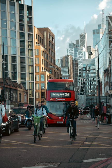 In the foreground, several pedestrians, including a man wearing sunglasses and a dark jacket, are crossing the street either on bicycles or walking, with their movement suggesting typical urban activity. To the left, a row of parked cars is visible along the curb, with sleek, modern vehicle designs and dark finishes reflecting the surrounding cityscape. Behind them, a red double-decker bus patterned with route information displaying 'Hackney Wick 26' is approaching or waiting at a traffic signal, its bright color standing out amid the urban environment. The background features a dense cluster of tall, glass-fronted office buildings and high-rise towers, some with reflective surfaces that mirror the partly cloudy sky above. The buildings exhibit contemporary architectural styles, with angles, setbacks, and a mixture of materials such as glass, steel, and brick, creating a dynamic city skyline backdrop. The scene is illuminated by natural daylight, casting soft shadows and highlighting the textures of the various buildings, vehicles, and street furniture, capturing a typical busy day in central Hackney with a focus on mobility and urban infrastructure, relevant to the context of alternative waste management and rubbish removal services like those offered by Waste Disposal Hackney.
