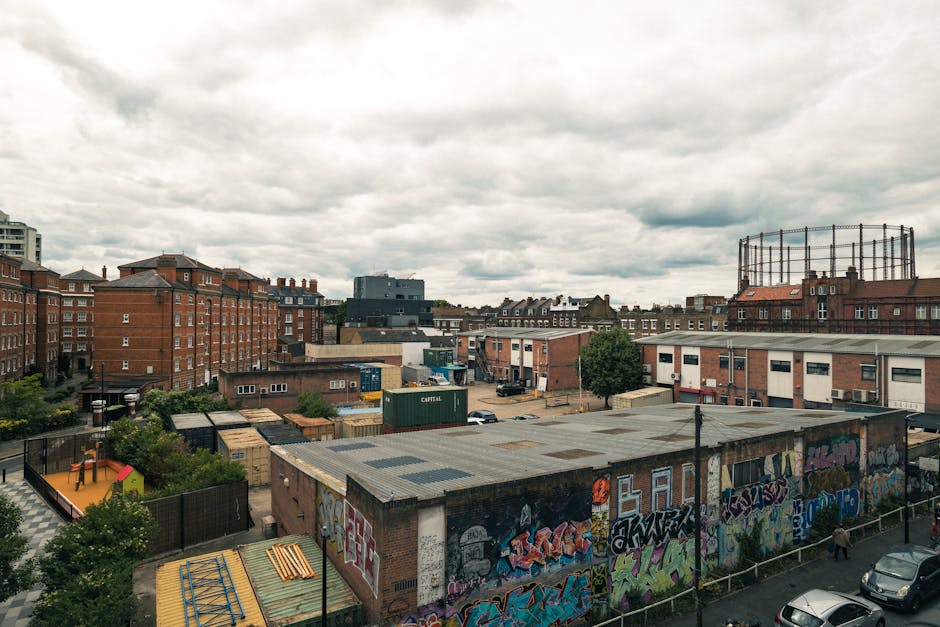 The image depicts an urban area with a view of various brick and concrete buildings, including residential apartments with red brick facades and flat roofs, as well as industrial or commercial structures with metal roofing. In the foreground, there is a single-story building covered in colorful graffiti artwork, with a slightly weathered appearance and a flat, corrugated metal roof. Surrounding this building are small trees, bushes, and a fenced playground area with a yellow and red play structure. Several shipping containers and parked vehicles are visible in the background, indicating a mixed-use environment that includes storage and parking spaces. To the right, a large, circular metal framework suggests an industrial installation or water tower. The scene is cast under an overcast sky with thick, grey clouds, creating a subdued, diffused natural lighting across the scene, which is typical in urban environments where private waste disposal and rubbish removal services might be employed to manage waste from commercial and residential buildings, as could be handled by Waste Disposal Hackney.