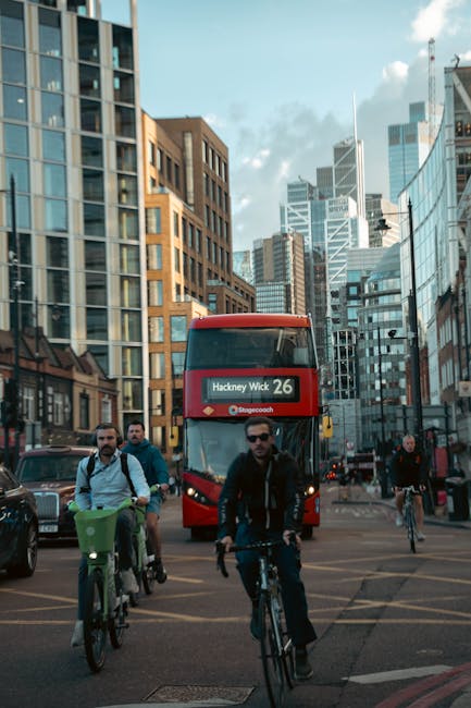 In the foreground, several pedestrians, including a man wearing sunglasses and a dark jacket, are crossing the street either on bicycles or walking, with their movement suggesting typical urban activity. To the left, a row of parked cars is visible along the curb, with sleek, modern vehicle designs and dark finishes reflecting the surrounding cityscape. Behind them, a red double-decker bus patterned with route information displaying 'Hackney Wick 26' is approaching or waiting at a traffic signal, its bright color standing out amid the urban environment. The background features a dense cluster of tall, glass-fronted office buildings and high-rise towers, some with reflective surfaces that mirror the partly cloudy sky above. The buildings exhibit contemporary architectural styles, with angles, setbacks, and a mixture of materials such as glass, steel, and brick, creating a dynamic city skyline backdrop. The scene is illuminated by natural daylight, casting soft shadows and highlighting the textures of the various buildings, vehicles, and street furniture, capturing a typical busy day in central Hackney with a focus on mobility and urban infrastructure, relevant to the context of alternative waste management and rubbish removal services like those offered by Waste Disposal Hackney.