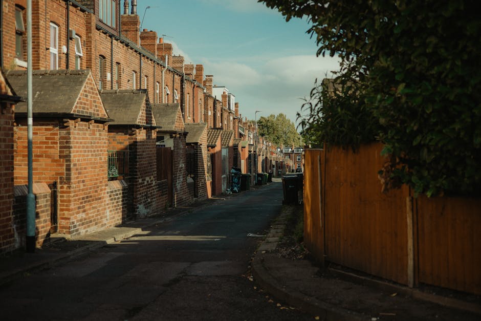 A narrow residential street lined with red-brick terraced houses featuring small front yards and pitched tiled roofs, some with chimneys visible. The pavement runs along the left side, with a concrete curb and a slight slope leading onto the street surface, which is paved with asphalt. On the right side, there is a tall wooden fence with a warm, natural finish, partially obscuring greenery from trees or bushes behind it. Several wheelie bins in dark colours are positioned along the curb, indicating waste collection points, and some houses have small garden elements visible through windows or near entrances. The street is illuminated by natural daylight, casting soft shadows on both the brickwork and the pavement, creating a calm, everyday suburban atmosphere. This scene reflects a typical inner-city residential area where private rubbish storage and disposal arrangements may be necessary, which companies like Waste Disposal Hackney can assist with through independent waste collection services suited to house clearance or ongoing rubbish removal needs.