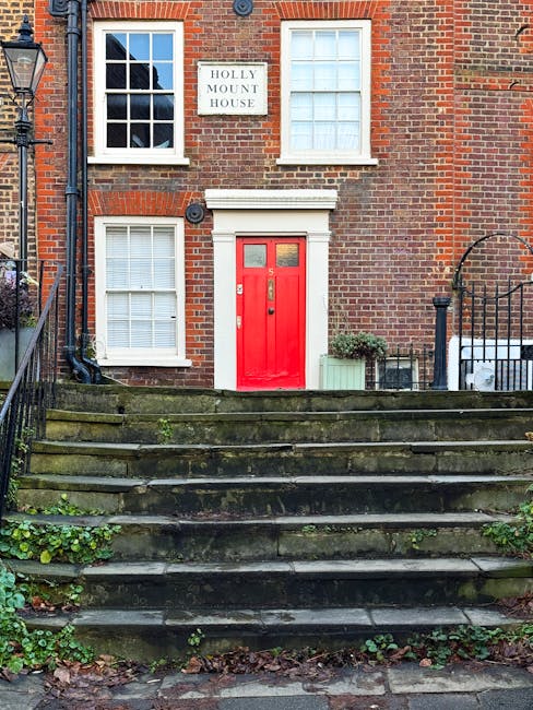 The image shows the front of a traditional brick residential building with a prominent bright red front door, framed by white trim, positioned centrally on a set of outdoor stone steps that are weathered and covered with patches of moss and small plants growing between the cracks. To the left of the door, there are two white-framed sash windows with closed blinds, one on the ground floor and one directly above on the first floor, both with a grid pattern dividing the glass panes. A white sign reading 'Holly Mount House' is mounted on the brick wall above and between the upper windows. To the right of the entrance, there is a small green potted plant and a light green planter box with greenery, placed on the landing area at the top of the steps. The surrounding environment includes a black iron railing on the right side of the stairs, supporting a small decorative gate, and a black street lamp post on the left side of the image. The scene is illuminated with natural daylight, highlighting the brickwork and details of the entrance, and creating a subtle contrast with the weathered stairs, which suggest a typical setting where private waste collection services might be employed instead of municipal rubbish collection.