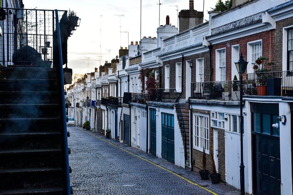 A narrow residential street lined with red-brick terraced houses featuring small front yards and pitched tiled roofs, some with chimneys visible. The pavement runs along the left side, with a concrete curb and a slight slope leading onto the street surface, which is paved with asphalt. On the right side, there is a tall wooden fence with a warm, natural finish, partially obscuring greenery from trees or bushes behind it. Several wheelie bins in dark colours are positioned along the curb, indicating waste collection points, and some houses have small garden elements visible through windows or near entrances. The street is illuminated by natural daylight, casting soft shadows on both the brickwork and the pavement, creating a calm, everyday suburban atmosphere. This scene reflects a typical inner-city residential area where private rubbish storage and disposal arrangements may be necessary, which companies like Waste Disposal Hackney can assist with through independent waste collection services suited to house clearance or ongoing rubbish removal needs.