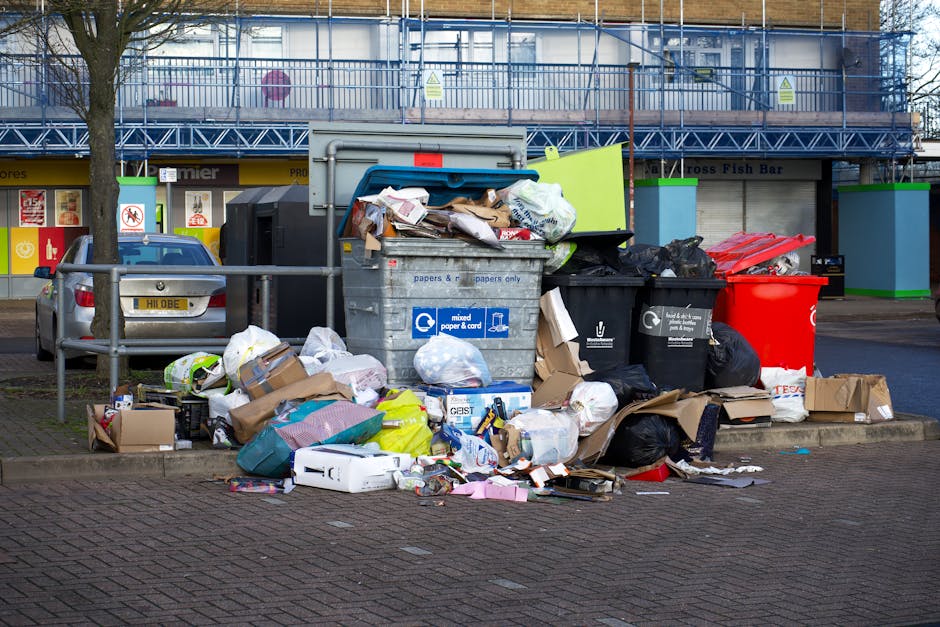 A collection of overflowing waste bins and scattered rubbish on a paved urban sidewalk in front of a commercial building, including a large grey mixed waste bin filled with paper, cardboard, and plastic waste, partially open with waste spilling out. To the right, there are black and red recycling bins, with black bags piled next to them containing additional waste, some tied and others loose. Surrounding the bins, various discarded items such as flattened cardboard boxes, paper packaging, plastic bags, and small debris are spread across the paved surface. In the background, a building with a blue protective metal scaffolding and a shop with a yellow sign is visible, alongside a parked grey car behind a metal railing, indicating a typical city centre environment. The scene reflects off-hours waste collection or excess rubbish awaiting removal, illustrating typical urban rubbish accumulation handled by private waste disposal services like Waste Disposal Hackney, in the context of alternative waste handling options.