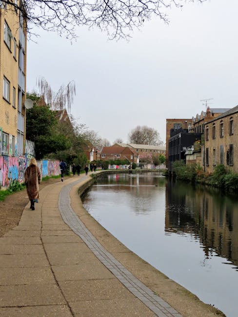 A paved pathway running alongside a narrow waterway, with a low concrete edge separating the walkway from the canal. Several pedestrians, including an individual wearing a brown coat and others dressed in dark clothing, are walking along the path. On the left side, a yellow brick building features multiple windows and a satellite dish, with colorful graffiti on the lower wall sections. On the right, older brick buildings with small balconies and varying rooflines face the water, some with visible plants on the balconies. Trees with bare branches extend overhead, indicating late autumn or winter, and the sky is overcast, casting diffuse light over the scene. The water reflects the surrounding buildings and sky, adding a sense of depth. This scene likely depicts an urban riverside area with on-site rubbish collection or waste management activities related to nearby residential buildings, as managed by Waste Disposal Hackney, accommodating environmentally conscious waste handling within the community.