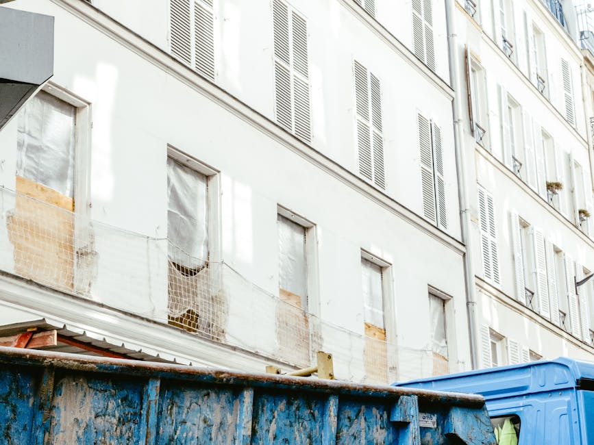 A large, blue Industrial waste skip with visible rust and dirt on its surface is positioned on a city street near a multi-storey residential building. The skip is partially filled with assorted construction and domestic waste, including cardboard boxes, plastic wrapping, and discarded materials, indicating an ongoing on-site clearance or renovation disposal activity. The surrounding environment includes the building's white facade with multiple closed wooden shutters painted in pale tones, some partially covered with protective netting for window repairs. The building’s exterior shows signs of weathering, with patches of brickwork exposed beneath peeling paint. The scene is illuminated by natural daylight, casting soft shadows and highlighting the textures of the skip, waste, and building surfaces. The presence of the skip suggests private waste collection services, such as those offered by Waste Disposal Hackney, as an alternative to standard council rubbish collection for larger or builder’s waste. This setup aligns with practices for managing non-standard rubbish removal tasks within urban environments, emphasizing the importance of proper waste handling for property maintenance or renovation projects.