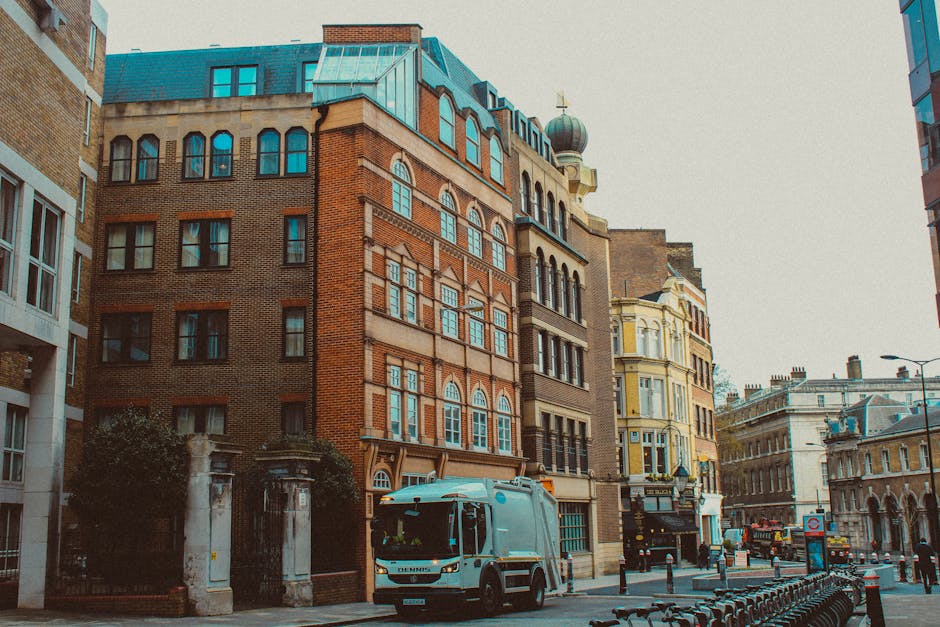 A white waste collection truck from Waste Disposal Hackney is parked on a city street bordered by a mix of historic and modern buildings with distinctive architectural features. The truck is positioned in the foreground, facing left, with its cab and cargo area clearly visible; it has a smooth, metallic finish and streamlined design. To the left, there is a wrought-iron gate supported by stone pillars, leading to an entrance of a building partially obscured by trees and shrubbery. The surrounding buildings display various materials and finishes, including brickwork and painted facades, in shades of brown, beige, and pale yellow, with multiple levels characterized by large windows, some with arched tops, and ornate decorative elements such as cornices and moldings. The street surface appears to be asphalt, with a row of metal bike racks and bollards along the sidewalk. Overhead, a cloudy sky diffuses natural light, creating a neutral, evenly lit scene that aligns with the context of private waste handling and alternative rubbish collection methods often represented by independent waste collection services like Waste Disposal Hackney.