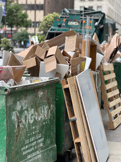 A large green waste disposal bin filled with a variety of discarded cardboard boxes, some flattened and others still in three-dimensional form, along with pieces of paper packaging. The boxes are made of brown corrugated cardboard, with some exhibiting printed labels or tape remnants. The bin, showing signs of weathering with chipped paint and dirt, is situated on an urban street with a backdrop of a cityscape that includes trees, multi-story buildings, and a parked dark green garbage truck with visible lights and equipment. To the right, there is a wooden pallet leaning against the bin, partially obscured by the boxes. The scene is illuminated by natural daylight, creating a clear, neutral atmosphere appropriate for descriptions related to independent waste collection or private rubbish disposal services, illustrating the process of clearing waste for alternative rubbish removal methods such as those offered by Waste Disposal Hackney.