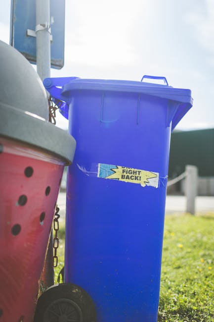 A large green waste disposal bin filled with a variety of discarded cardboard boxes, some flattened and others still in three-dimensional form, along with pieces of paper packaging. The boxes are made of brown corrugated cardboard, with some exhibiting printed labels or tape remnants. The bin, showing signs of weathering with chipped paint and dirt, is situated on an urban street with a backdrop of a cityscape that includes trees, multi-story buildings, and a parked dark green garbage truck with visible lights and equipment. To the right, there is a wooden pallet leaning against the bin, partially obscured by the boxes. The scene is illuminated by natural daylight, creating a clear, neutral atmosphere appropriate for descriptions related to independent waste collection or private rubbish disposal services, illustrating the process of clearing waste for alternative rubbish removal methods such as those offered by Waste Disposal Hackney.