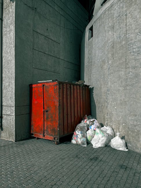 A large pile of mixed waste materials, consisting of cardboard boxes, plastic bags, paper, and various household items, is overflowing from multiple rubbish bins and scattered on the paved surface of a parking area. The scene includes a grey wheeled bin positioned in the center, filled with crumpled paper, cardboard, and other refuse, with some waste spilling onto the ground. Surrounding the bin are black, red, and beige plastic bags filled with waste, as well as flattened cardboard boxes and loose paper. To the left of the waste pile, there is a parked silver car with a UK registration plate, situated behind a metal railing that separates the sidewalk from the parking area. In the background, a commercial building with a blue scaffolding structure and a row of shops, including a shop with a yellow sign, can be seen, indicating an urban environment. The scene appears to be a typical example of improperly managed rubbish accumulation, relevant to private waste disposal or alternative collection services provided by waste management companies like Waste Disposal Hackney, emphasizing the importance of effective rubbish removal and disposal methods.